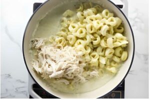 Pot of cheese tortellini, shredded chicken, and creamy white sauce on stove.