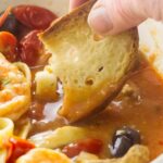 Hand dips crusty bread into shrimp, pasta, and tomato.