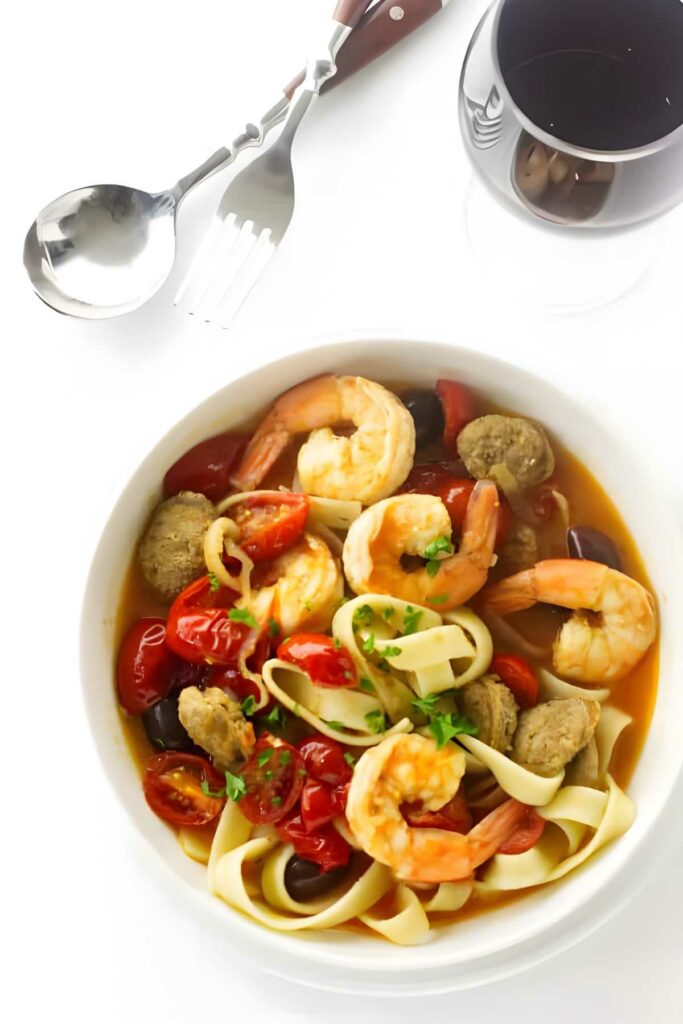 Pasta with shrimp, chorizo, and tomatoes beside red wine on white background.