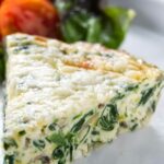 Slice of spinach and cheese quiche on white plate with greens and tomatoes.