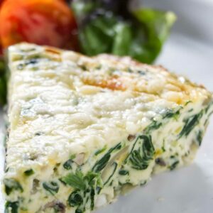 Slice of spinach and cheese quiche on white plate with greens and tomatoes.