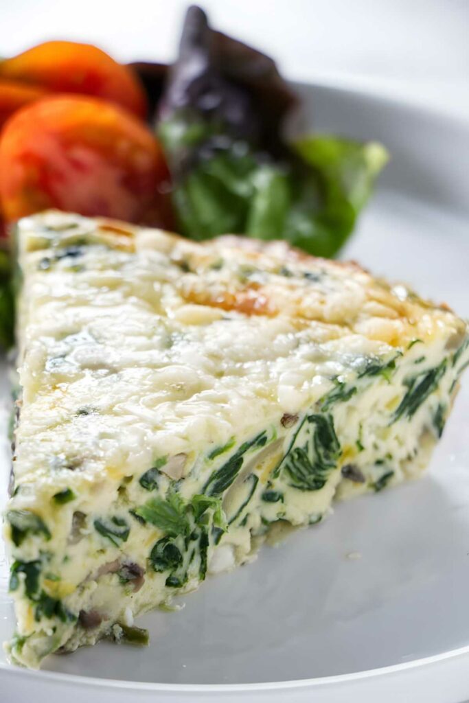 Slice of spinach and cheese quiche on white plate with greens and tomatoes.