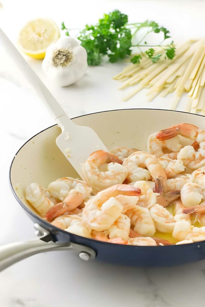 Shrimp sautéing in a skillet with white spatula; pasta, garlic, lemon nearby.