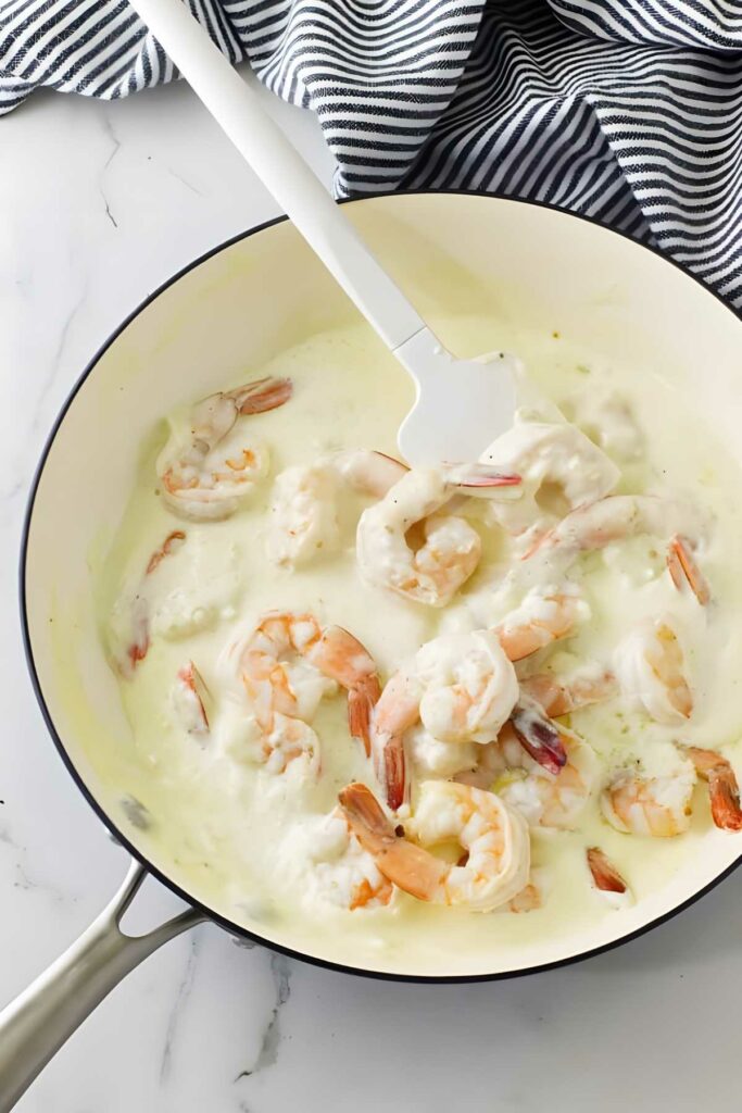 Shrimp cooking in creamy sauce in white skillet, stirred with white spatula.