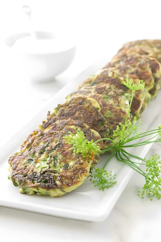 Golden-brown zucchini fritters with herbs on a white rectangular plate.