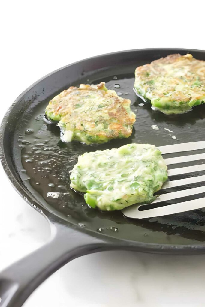 Three green vegetable fritters cooking in skillet, one lifted to show golden side.