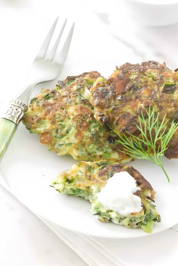Three golden-brown vegetable fritters with sour cream and dill on a white plate.