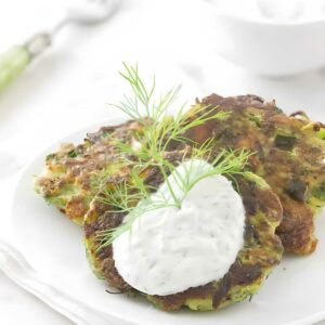 Two zucchini fritters with herb sauce and dill on a white plate.