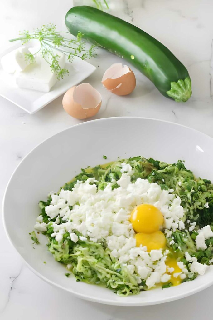White bowl with shredded zucchini, feta, raw eggs; ingredients nearby on marble.