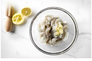 Glass bowl with raw shrimp and garlic, lemon halves and citrus juicer nearby.