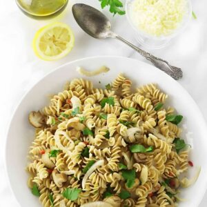 Bowl of rotini pasta with mushrooms, parsley, and red pepper on white surface.