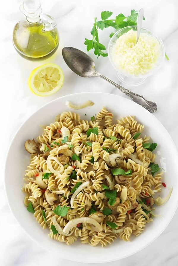 Bowl of rotini pasta with mushrooms, parsley, and red pepper on white surface.
