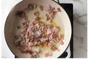 Chopped bacon cooking in a white pan, some browned, some still pink.
