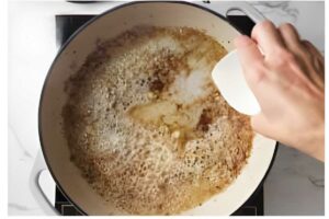 Hand scrapes browned bits from white pot with spatula on stovetop.