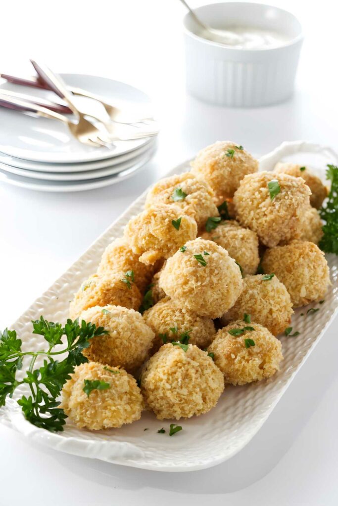 Golden baked balls with parsley on a white platter, dipping sauce behind.