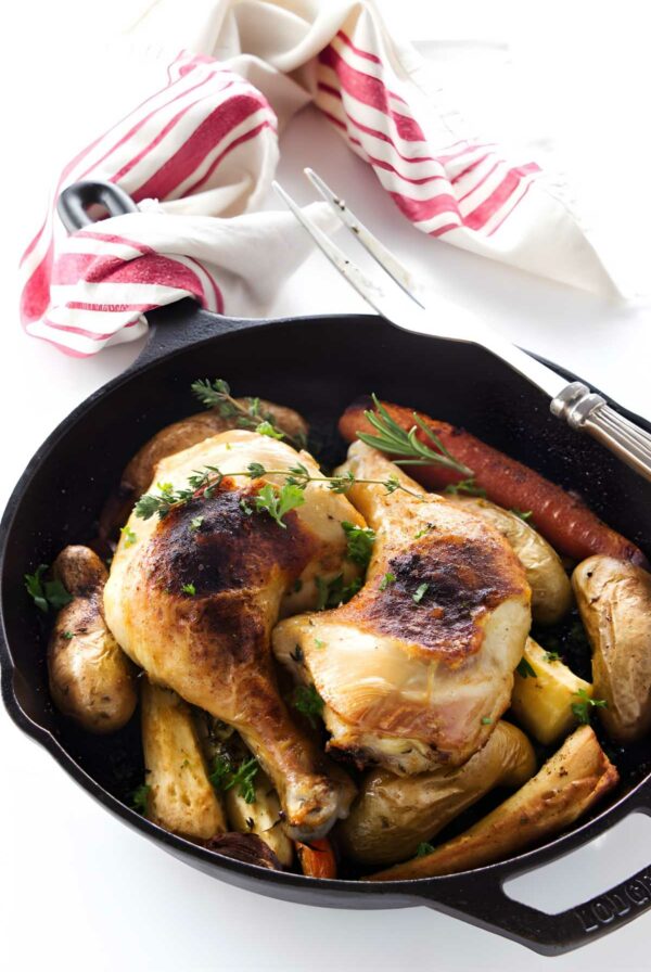 Cast iron skillet with roasted chicken, root vegetables, and herbs, on white surface.