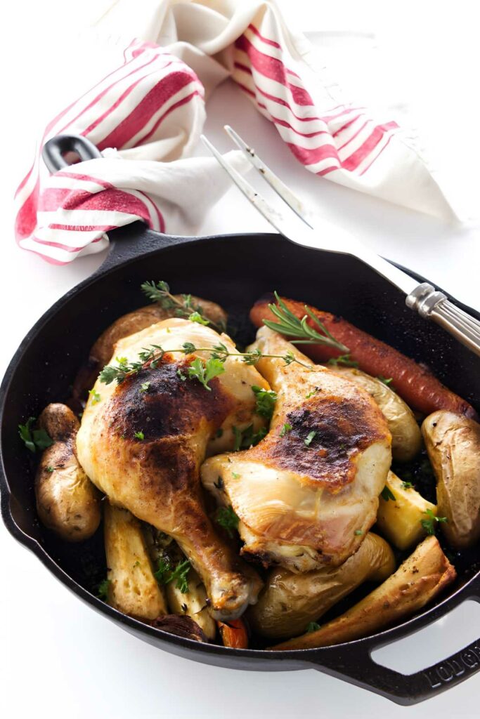 Cast iron skillet with roasted chicken, root vegetables, and herbs, on white surface.