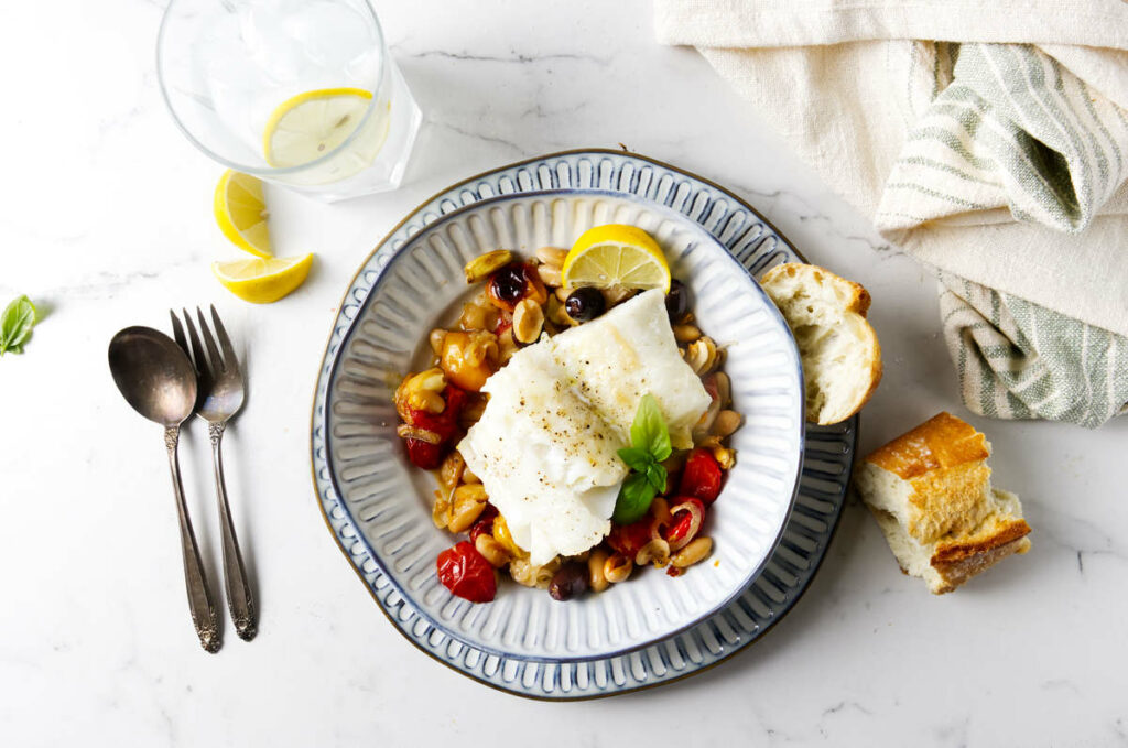 A bowl with a serving of roasted cod with tomatoes and beans next to a slice of bread.