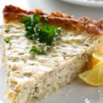 Close-up of a quiche slice with parsley and lemon wedges on a plate.