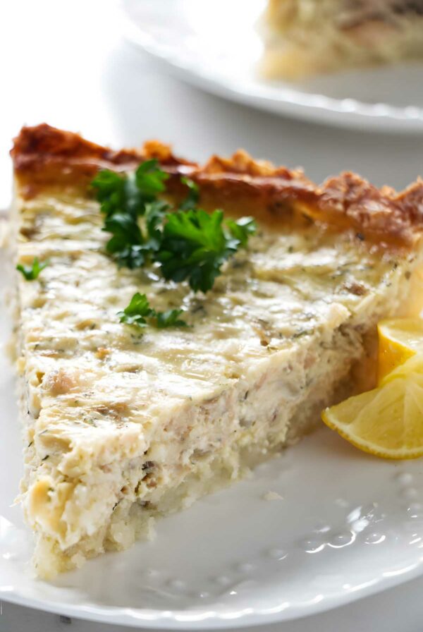 Close-up of a quiche slice with parsley and lemon wedges on a plate.