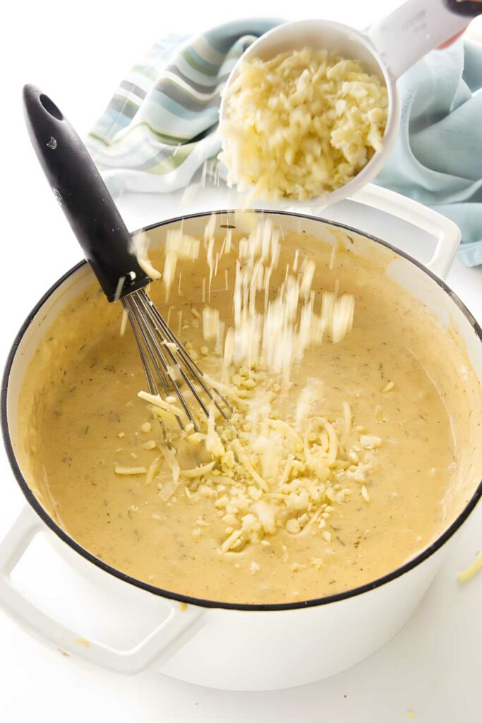 Cheese poured from a cup into creamy sauce, whisk inside pot.