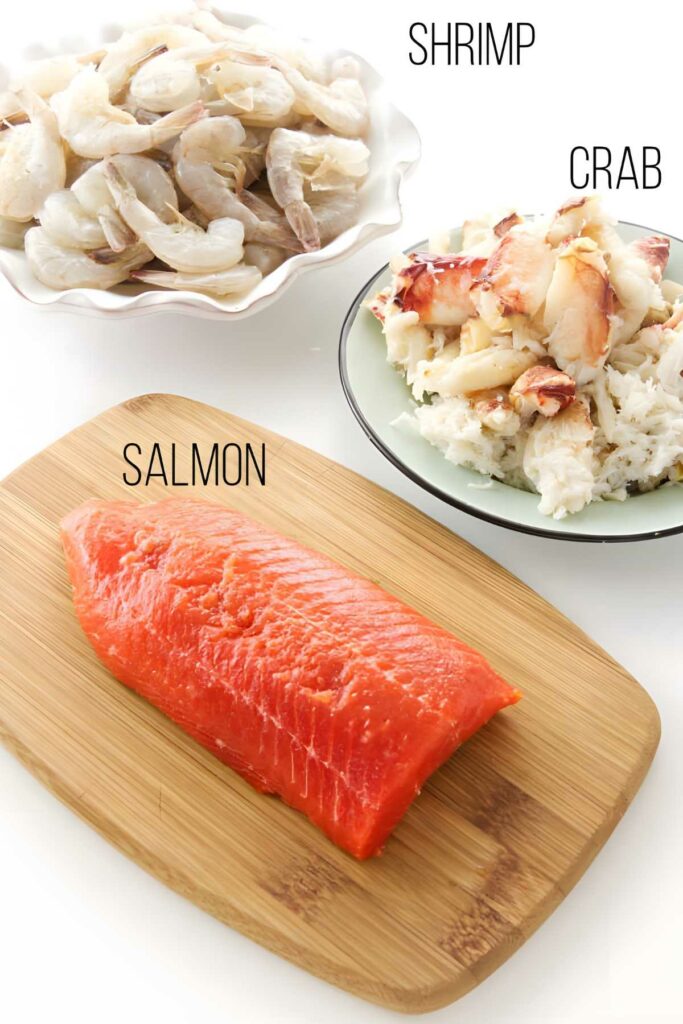 Salmon fillet, shrimp and crab on a wooden board, plates of raw shrimp and crab meat nearby.
