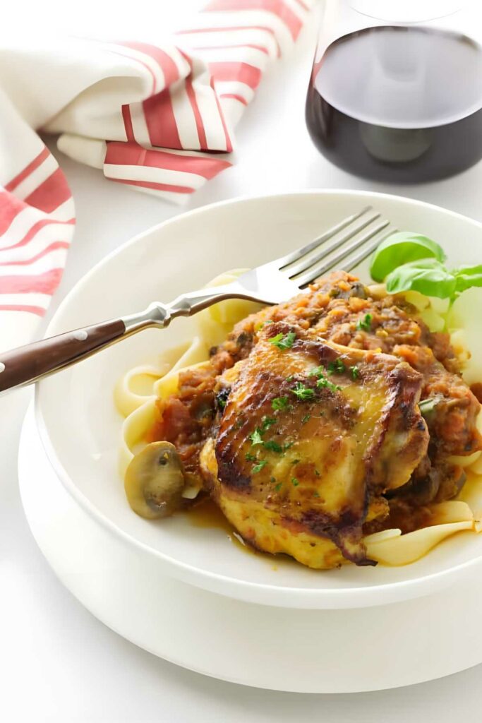 Pasta with braised chicken and vegetables, garnished, beside red wine and utensils.