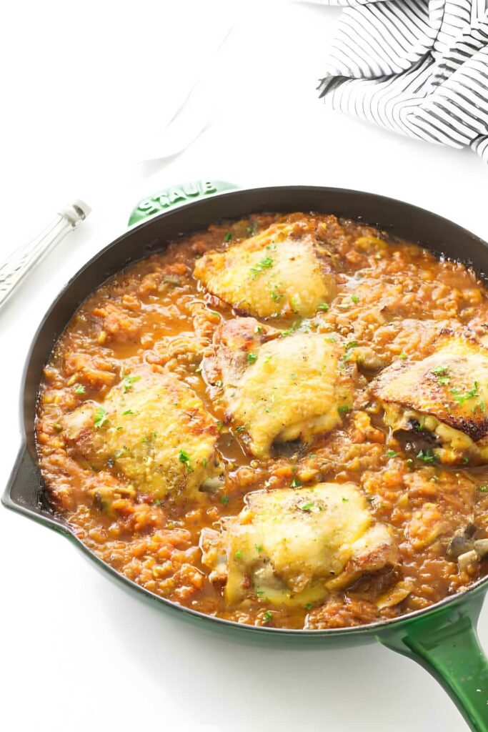 Chicken thighs in tomato sauce with herbs, in skillet on white surface.