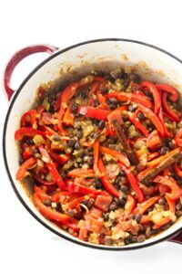 Red skillet with cooked red peppers, tomatoes, and onions, top view.