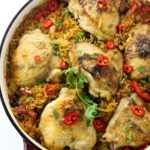 Golden-brown chicken thighs on rice in a red Dutch oven, garnished with chili and cilantro.