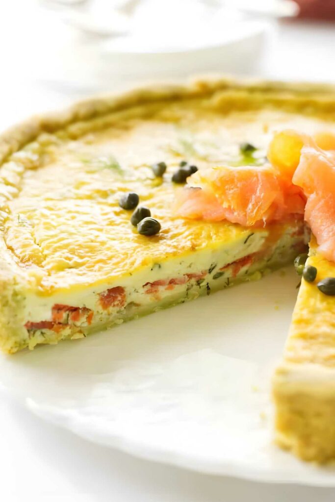 Close-up of sliced quiche with smoked salmon, capers, herbs on white plate.