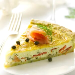 Quiche slice with smoked salmon, capers, and dill on white plate.