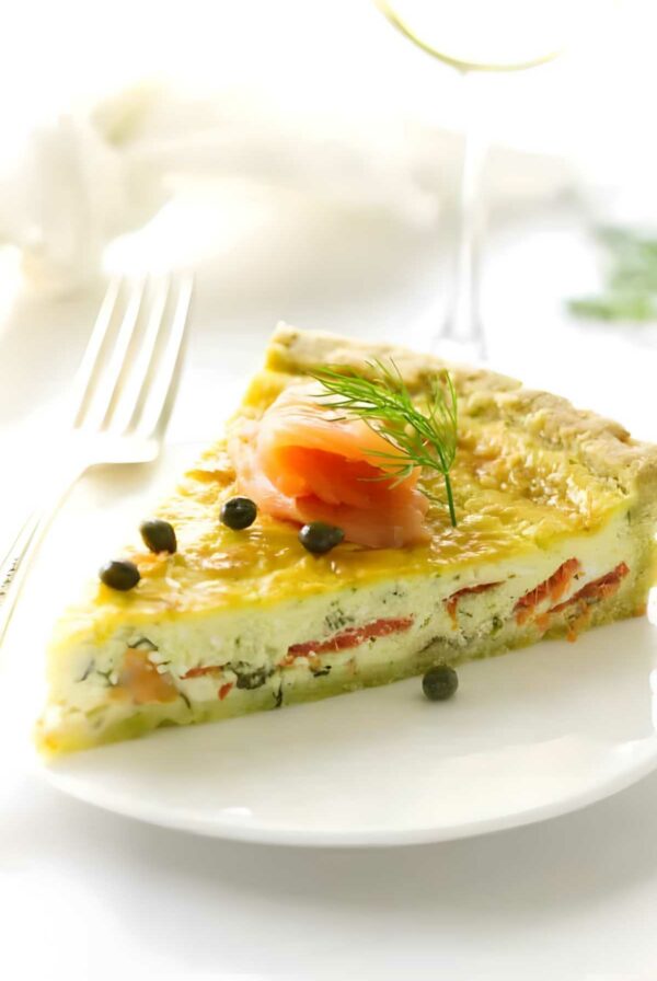 Quiche slice with smoked salmon, capers, and dill on white plate.