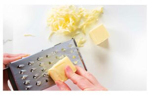 Hand grating butter with a box grater on a white surface.