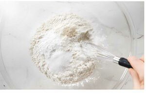 Hand whisking dry baking ingredients in a clear glass bowl on white surface.