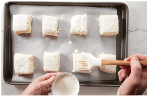Brushing liquid onto unbaked biscuit dough squares on a baking sheet.