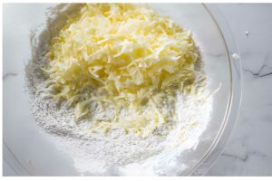 Glass bowl with grated cheese atop flour, on white marble surface.