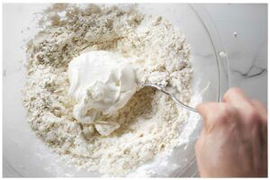 Hand mixes yogurt into flour in a bowl on a white countertop.