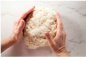 Hands kneading crumbly dough on white marble surface.