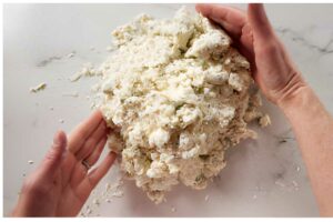 Hands shaping crumbly dough on a floured white countertop.