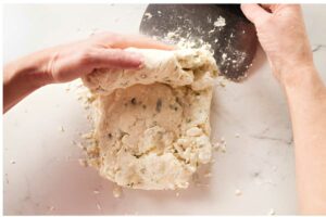 Hands shaping crumbly dough on white surface with metal bench scraper.