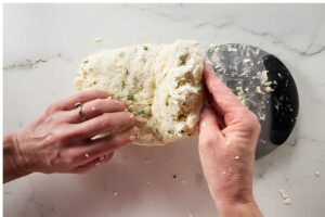 Hands knead crumbly dough on marble beside a dough-covered metal scraper.