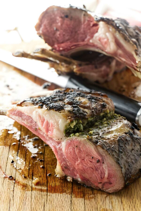 Sliced rack of lamb with herb stuffing on a wooden cutting board, knife nearby.