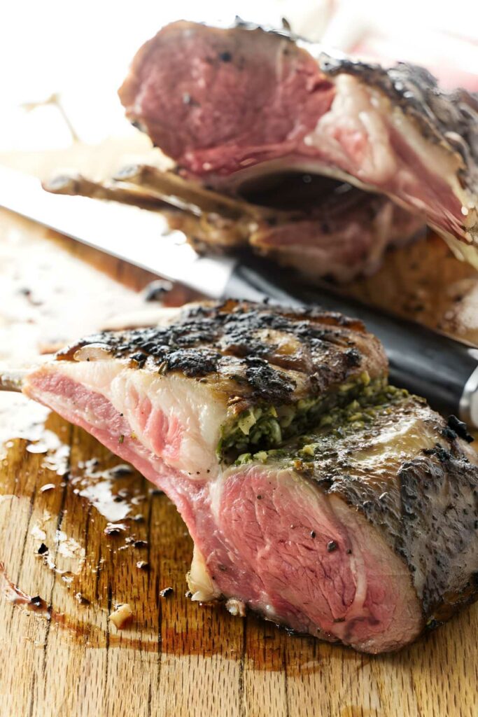 Sliced rack of lamb with herb stuffing on a wooden cutting board, knife nearby.