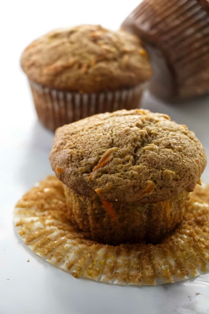 Close-up of a spelt muffin with liner peeled back, two blurred muffins behind.
