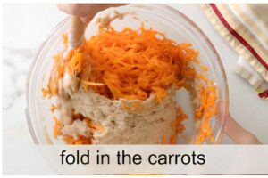 Hand folding shredded carrots into batter with spatula; text reads fold in the carrots.