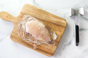 A chicken breast on a wood board, wrapped in plastic next to a meat mallet.