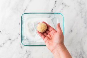 A hand rolling sugar cookie dough in granulated sugar.