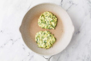 Two zucchini fritters cooking in a skillet.