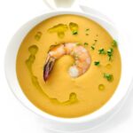 Bowl of orange soup with shrimp, herbs, olive oil, and a spoon.